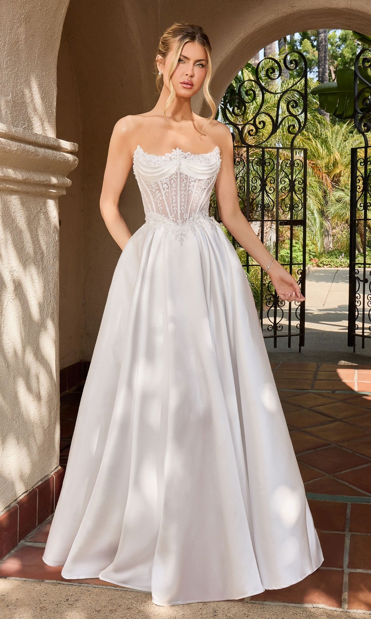 Wearing the Ladivine CC511W, a white strapless formal dress with an embroidered corset bodice and flowing A-line silhouette, a woman stands gracefully under an outdoor archway.